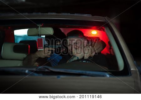 Car thief threatening driver with the gun. Male robber wearing black mask sitting at the back seat and pointing gun at the head of scared owner. Police light on the background.