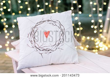 indoor, house, rest concept. on the background of beautiful magestic blurs there is white pillow with nice print in form of heart surrounded by wreath of brunches without leaves