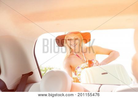 Happy woman loading suitcase in car trunk