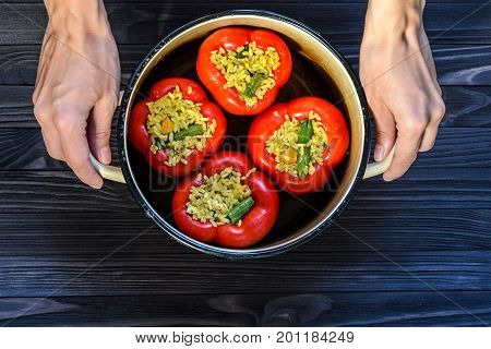 Chef holding a pan with four red stuffed peppers on dark rustic kitchen table background top view. Cooking process