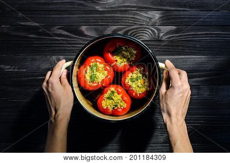 Chef holding a pan with four red stuffed peppers on dark rustic kitchen table background first person view. Cooking process top view