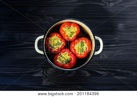 Four red stuffed peppers in a yellow saucepan on dark rustic kitchen table background top view. Cooking process