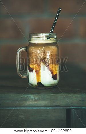 Iced caramel macciato coffee with milk in glass jar with straw on dark wooden table, selective focus. Cold sweet refreshing summer drink concept