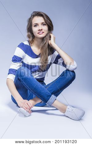 Trendy and Fashionable Positive Caucasian Brunette Girl Sitting on Floor With Crossed Legs and Touching Hair In Studio Environment.Vertical Image Orientation