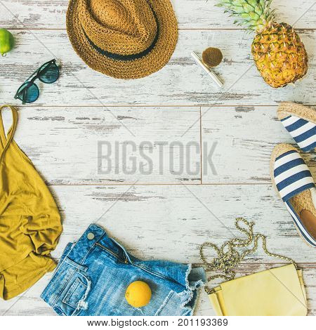 Colorful fashion outfit flat-lay. Denim shorts, straw hat, yellow top, espadrillas, leather bag, sunglasses, pineapple, limes, lemon over pastel parquet background, top view, square crop copy space