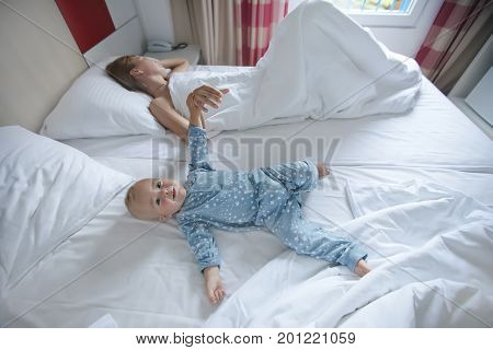 Adorable baby lying in bed with woman sleeping and holding baby's hand.