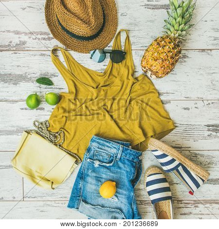 Colorful summer fashion outfit flat-lay. Denim shorts, straw sun hat, yellow top, espadrillas, leather bag, sunglasses, pineapple, limes and lemon over pastel parquet background, top view, square crop