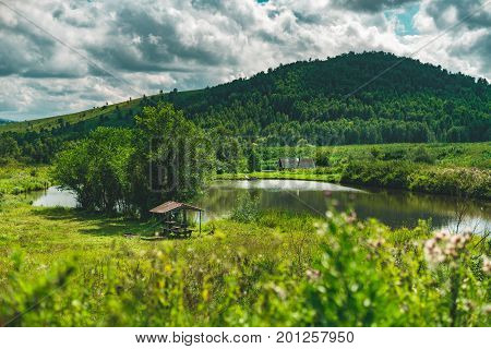 Summer landscape with small lake of fresh water hills with forests in distance green meadows of native grasses arbour with trees near submerged country house Altai mountains Aya district Russia