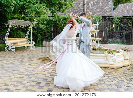 Happy groom and beautiful redhead bride in white dress holding hands and dancing outdoors free space. Beautiful wedding couple spinning in wedding day. Wedding dress with bow on back flutters in the wind