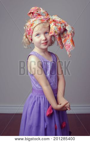 Portrait of cute adorable white blonde Caucasian preschool girl in purple dress with large bow on her head. Child posing in studio home on light plain background. Kid having fun. Toned with filters.