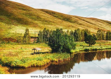 Autumn scenery with small lake of fresh water hills in distance green and yellow meadows of native grasses abandoned arbour with trees near Altai mountains Aya district Russia
