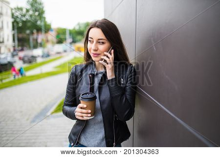 Beautiful Student Girl Leaning On Wall, Talking On Smart Phone And Holding Takeaway Coffee In Paperc