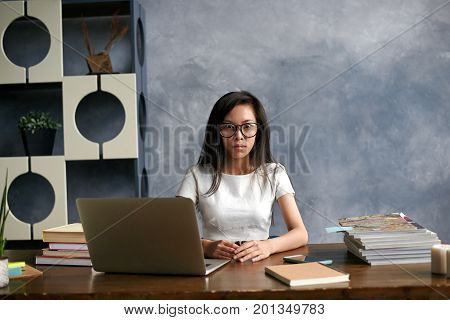Picture of shocked Asian student girl wearing glasses sitting at desk with laptop and piles of books looking puzzled as she has to do a lot of homework within a short time looking at camera in shock