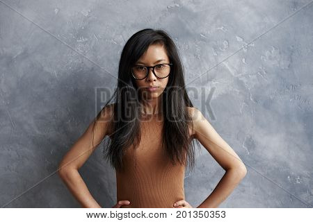 Studio shot of annoyed irritated brunette girl of Asian appearance wearing eyeglasses posing at grey wall with hands on her waist feeling mad and furious about something hel look full of anger