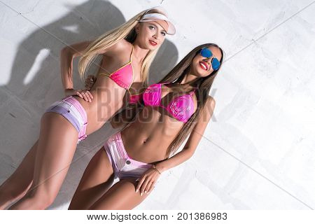 Sexy tanned girls wearing pink bicinis posing against white brick wall sunny shot