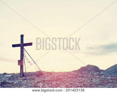 Modest Wooden Cross Raised On Rocky Alpine Mountain Summit . Sharp Rocky Peak. Gentle Clouds  In Blu