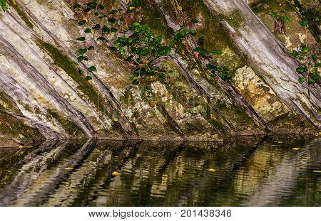 Monolithic wall of rock covered with moss and ivy reflected in the water of the lake river