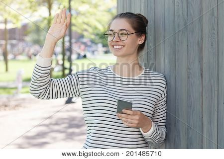 Outdoor Image Of Young Happy European Woman Greeting Mate She Sees In Street With One Hand And Holdi