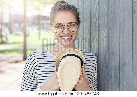 Closeup Photo Of Young Smiling European Female Leaning To Gray Wooden Wall With Shoulder, Holding Su