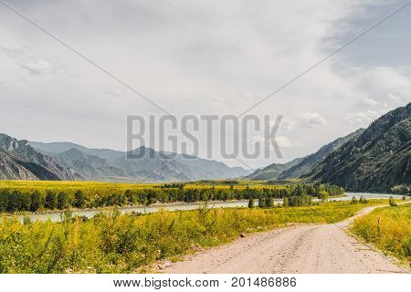 Dirt road stretching to distance with meadows of native grasses on sides mountain river Katun and ridges in hazy distance overcast sky Altai Kuyus district Russia