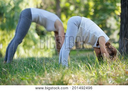 Young sporty mother and cute little daughter doing yoga exercises on grass in the park at the day time. Concept of friendly family and healthy lifestyle