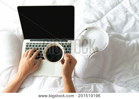 Male sitting on white bed surfing internet or working on laptop and listening to musical media online with wireless headphone and drink coffee in morning relax mood. Music and Lifestyle Concept.