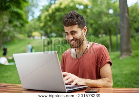People, beauty and lifestyle concept. Shot of handsome man with beard and wide smile dressed in red T-shirt smiling broadly being happy to receive e-mail with good news, typing reply to this e-mail. Attractive hipster sitting in front of monitor of his no