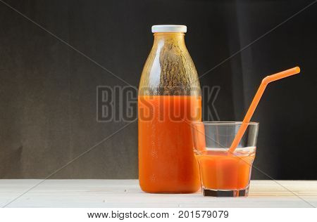 A bottle of carrot juice and a glass of carrot juice with a straw on white wood table on black background. Still life of a healthy eating concept