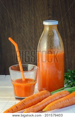 A bottle of carrot juice and a glass of carrot juice with a straw on white wood table against brown wood wall background. Still life of a healthy eating concept