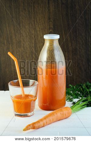 A bottle of carrot juice and a glass of carrot juice with a straw on white wood table against brown wood wall background. Still life of a healthy eating concept