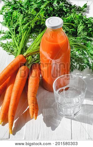 A bottle of carrot juice with empty glass and some fresh carrots on white wood table. Still life of a healthy eating concept