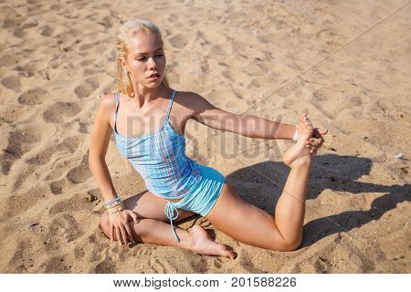 Blonde long haired girl in a blue suit is stretching and doing yoga on a beach in sunlight of the rising sun