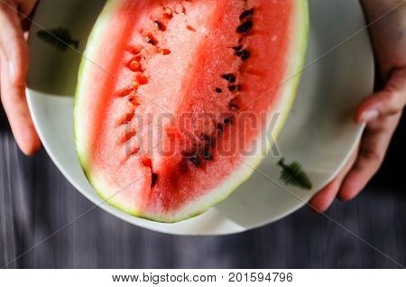 Slice of watermelon on white plate in woman hands ready for eating top view with blurry black wood table background