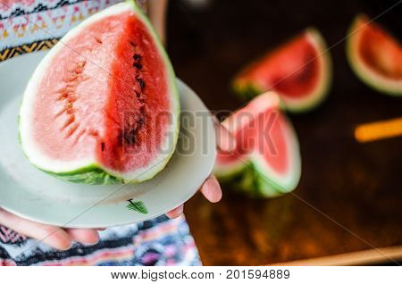 Watermelon background. Girl in a colorful summer shirt holding a plate with a large slice of watermelon against a background of a blurry table with slices of watermelon