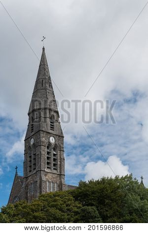 Clifden Ireland - August 4 2017: the gray spire of the local Catholic church tower against white clouds in blue sky. Some green of trees at bottom.