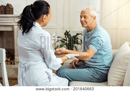 Try to relax. Pleasant nice skillful nurse sitting opposite her patient and holding his hand while preparing to do an injection