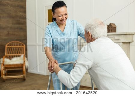 Friendly relationships. Positive cheerful nice woman looking at her patient and smiling while helping him to move around