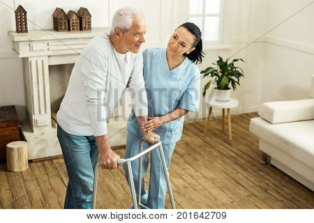 Professional aid. Pleasant positive senior man standing together with a caregiver and using walking equipment while moving around
