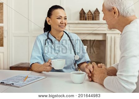 Pleasant drink. Cheerful delighted nice nurse holding a cup and having tea while talking to her elderly patient