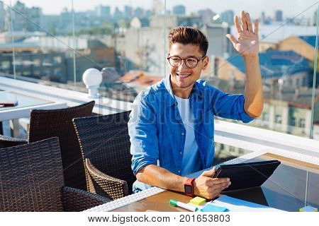 Hello there. Happy minded gentleman wearing glasses raising his hand and greeting someone at a cafe while relaxing on a terrace and using his tablet computer.