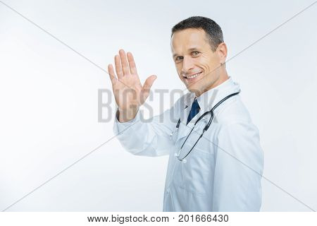 Hello there. Waist up shot of a friendly looking medical professional wearing a white coat saluting to a patient while standing over light background and grinning broadly into the camera.