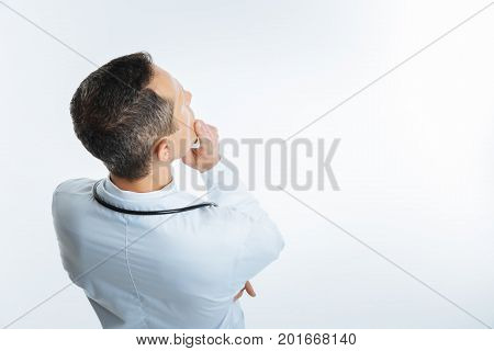 Hmmm. Turned back male physician wearing a white coat looking at an invisible screen and thinking of something alone over the light background.