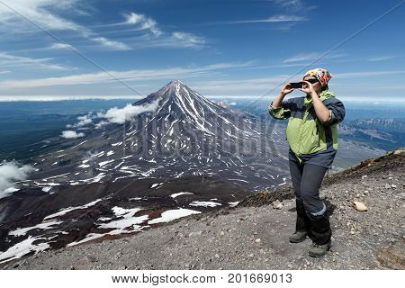 AVACHA VOLCANO KAMCHATKA PENINSULA RUSSIAN FAR EAST - AUG 7 2014: Women tourists standing on edge of crater of active Avacha Volcano and photographed on smartphone on background of Koryak Volcano.