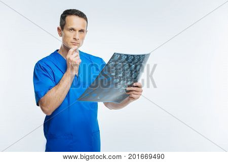 Hmmm. Waist up shot of a thoughtful medical professional touching his chin while standing with a mri scan of a patient head and thinking over it.