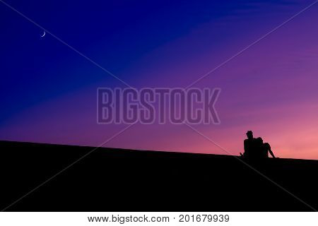 The silhouettes of two men sitting against the purple colors of a sunset with a slice of the moon on the blue part of the sky.