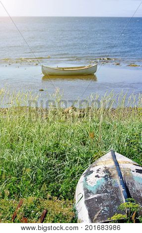 Old fishing boat lies upside down among the grass on a high seashore against the backdrop of the sea and moored in the bay by a stone ridge of a white wooden boat. Vertical frame
