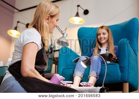 Pleasant chat. Pleasant young pedicurist removing the cuticle from toenails and talking to her client while the girl holding a magazine in her lap