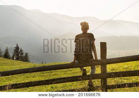 Young male tourist wih backpack sits on fence in hill side enjoying beautiful sight of mountains. Hiker takes rest and looks at distant ukrainian carpathian mountain range at sunset in summer