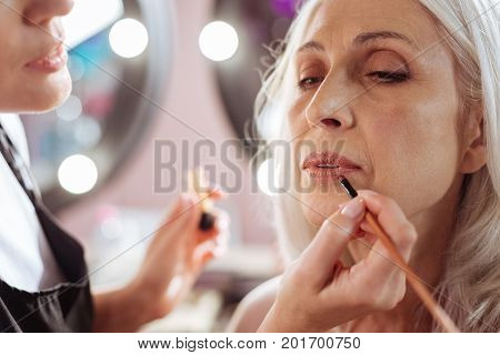 Soft lip care. The close up of delicate hands of a young makeup artist holding a makeup brush and applying lipstick to the lips of an elderly woman, finishing up her look