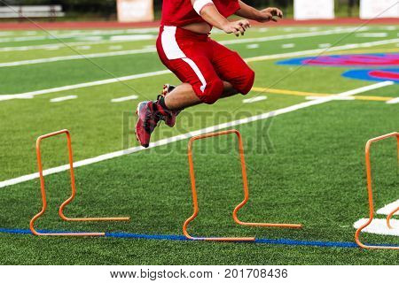 A football player is jumping over orange hurdles on a green turf field during high school practice on a warm August night.